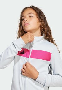 Persona joven con cabello rizado cerrándose una chaqueta blanca con una franja rosa y un logo negro en el pecho, frente a un fondo liso.