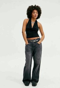 Top preto sem mangas com decote drapeado, combinado com jeans cinza escuro de corte largo e um cinto decorativo. Modelo está de pé com as mãos nos bolsos.