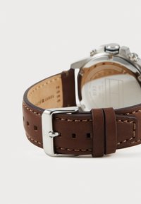 Bracelet de montre en cuir marron avec surpiqûres claires et boucle en métal argenté, présentant une finition lisse et un boîtier de montre circulaire visible par l'arrière.
