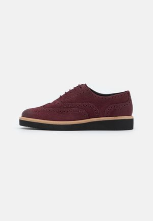 Chaussure brogue en daim rouge foncé avec des détails perforés, semelle intermédiaire beige et semelle en caoutchouc noire, vue de profil sur fond blanc.