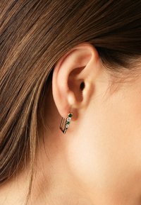 Orecchini a bottone in tono oro con cristalli verdi e trasparenti in un design quadrato. Accenti dorati texturizzati e lucidi sono presenti lungo i bordi.