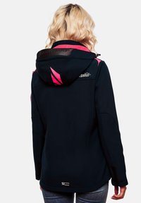 Woman with blonde wavy hair wearing a black and pink hooded jacket, standing facing away, with visible logos on the hood and back.