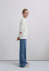 Kermainen collegepaita, jossa värikkäät tähtikuvioinnit hihoissa, yhdistettynä leveälahkeisiin sinisiin farkkuihin, joissa on tähtikirjailut saumoissa.