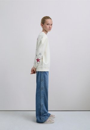 Bluză crem cu imprimeu colorat de stele pe mâneci, asortată cu blugi albastri cu croială largă, având broderie cu stele de-a lungul cusăturilor.