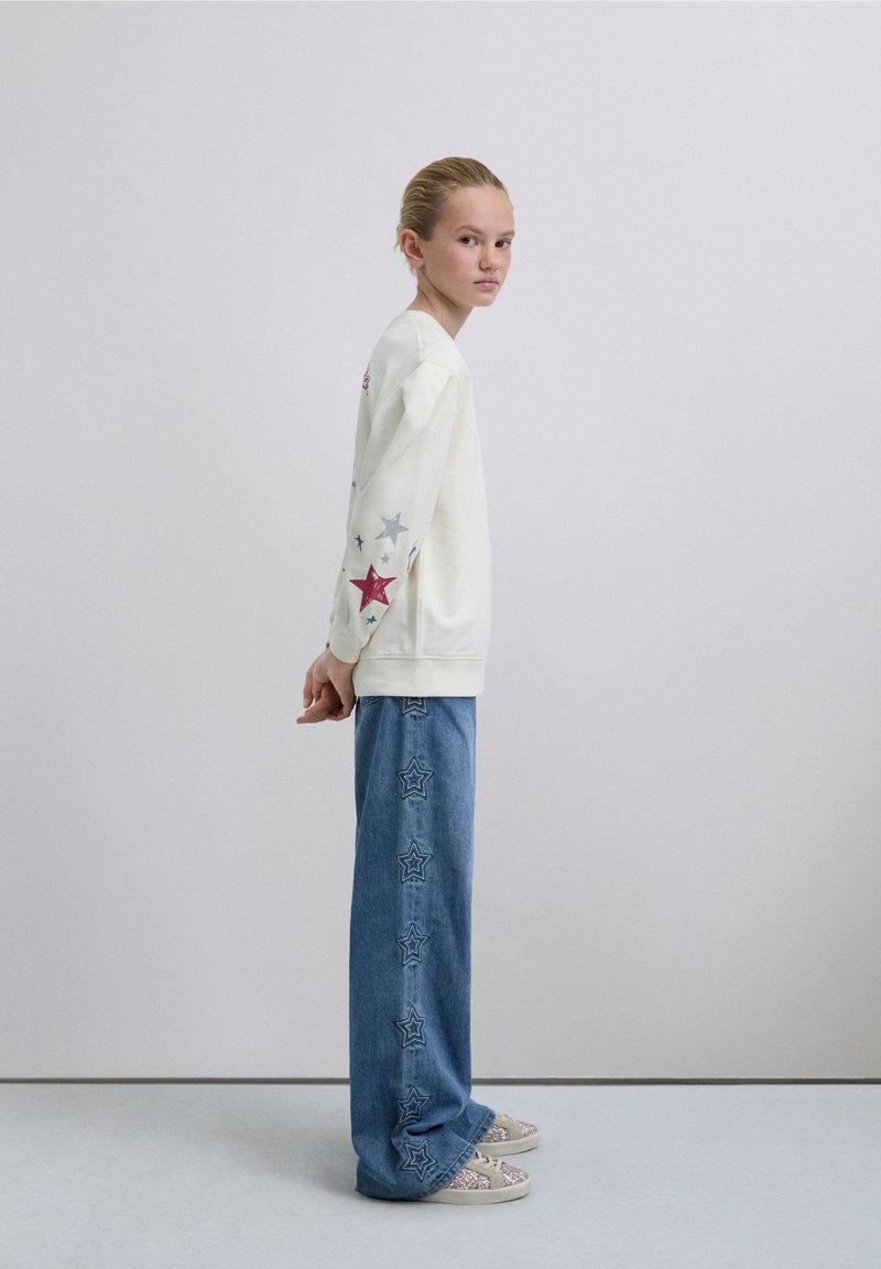 Kermainen collegepaita, jossa värikkäät tähtikuvioinnit hihoissa, yhdistettynä leveälahkeisiin sinisiin farkkuihin, joissa on tähtikirjailut saumoissa.