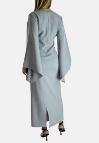 Lungo vestito grigio con una texture liscia, maniche a campana e una cerniera sul retro, caratterizzato da uno spacco laterale per facilitare i movimenti.