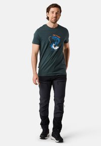 Mørkegrøn bomulds T-shirt med et fiske-motiv, kombineret med sorte lynlåse cargo-bukser og sorte sportssko, stående foran en neutral baggrund.