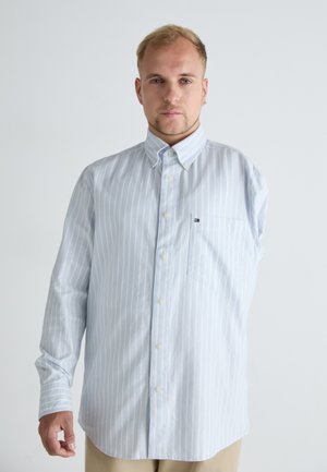 Jeune homme aux cheveux blonds courts portant une chemise à rayures bleu clair boutonnée, debout devant un fond blanc uni.
