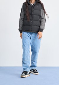 Person, der eine schwarze Steppweste über einem grauen Hoodie trägt, helle blaue Jeans mit großen Fronttaschen und schwarz-weiße Sneakers auf einem blauen Boden.