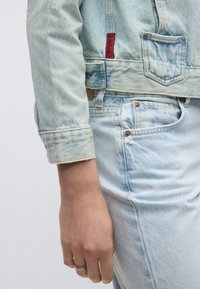 Ανοιχτό μπλε τζιν μπουφάν με ελαφρώς ξεθωριασμένη όψη, φέρει κόκκινη ετικέτα, μανίκια γυρισμένα προς τα πάνω και εμφανείς ραφές.