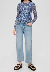 Kvinna klädd i en långärmad tröja med blå och beige mönster, ljusblå högmidjade raka jeans och svarta sandaler med öppna tår och remmar.