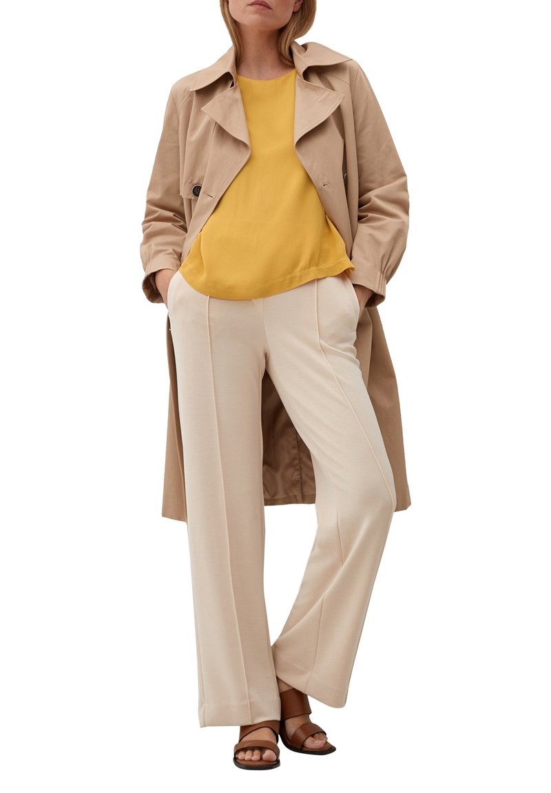 Femme portant un trench beige sur un chemisier moutarde et un pantalon large crème avec des sandales marron à bout ouvert, mains dans les poches, debout.