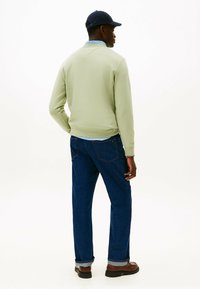Felpa verde in pile con polsini a costine, indossata sopra una camicia azzurro chiaro, abbinata a jeans in denim scuri e scarpe nere. Un berretto aggiunge un tocco di dettaglio.