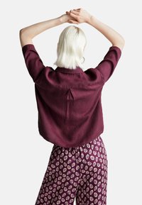 Persona con capelli platino biondi corti, che indossa una camicia larga di prugna scura e pantaloni con motivo, in piedi con le braccia sollevate e le mani unite dietro la testa.