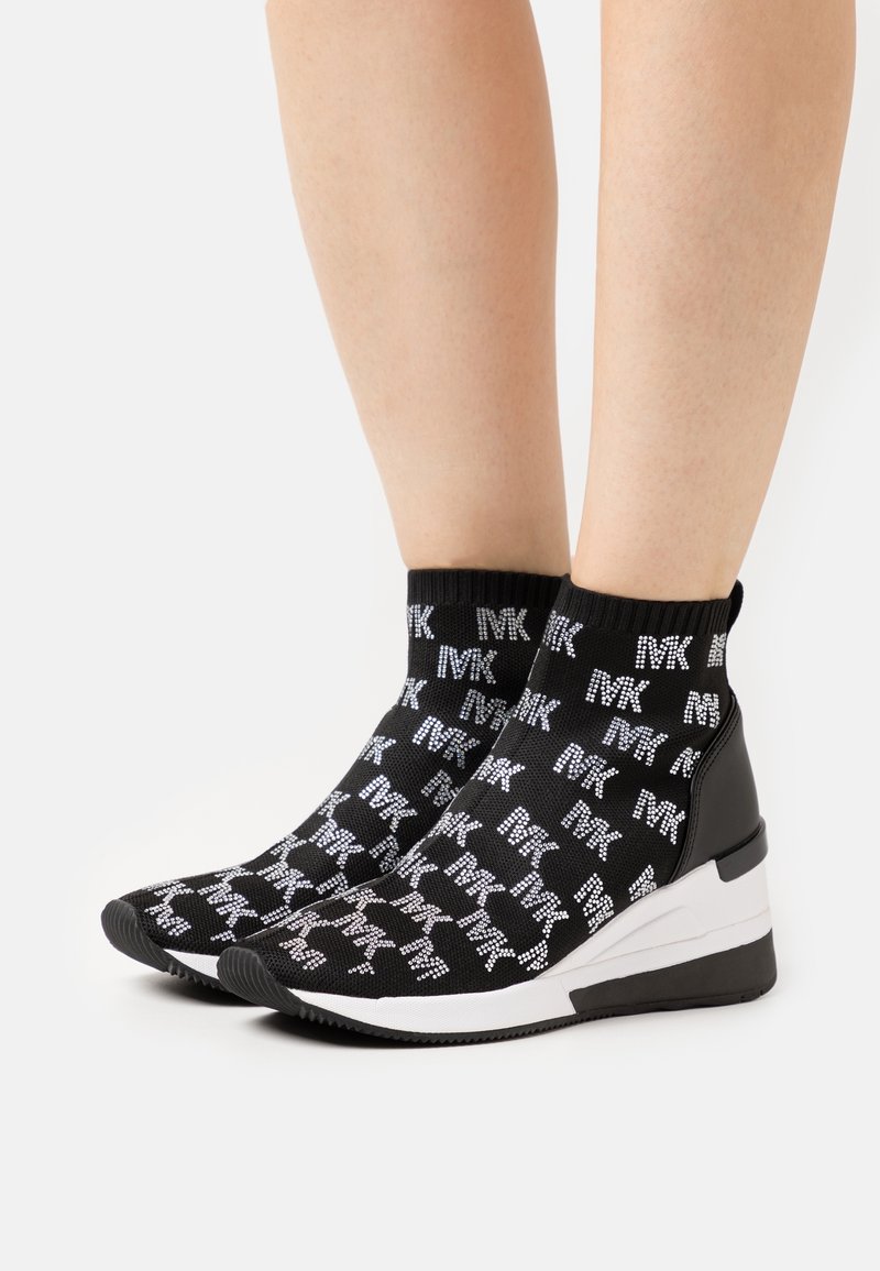Füße in schwarzen und weißen Keil-Sneakern mit einem strickähnlichen Obermaterial, das mit wiederholten "MK"-Logos gemustert ist.