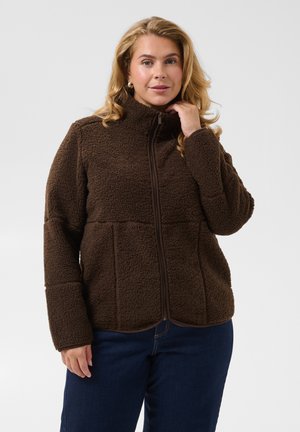 Femme aux cheveux blonds portant une veste polaire marron zippée et un jean bleu foncé, debout devant un fond clair uni.