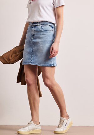 Kvinde iført lyseblåt denimskørt, hvide sneakers, hvid t-shirt, holder en brun jakke i hånden mod en ensfarvet baggrund.