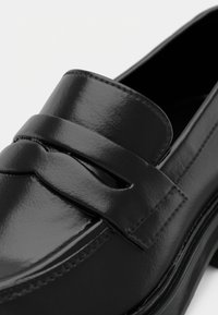 Gros plan d'un mocassin en cuir noir poli montrant les coutures et un détail de bride sur le dessus.