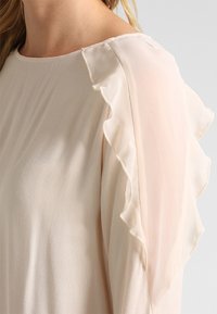 Top beige con un corpo testurizzato e maniche trasparenti a volant. Il tessuto sembra leggero e morbido, con una vestibilità rilassata attorno al collo.