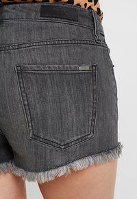 Pantalones cortos de mezclilla negra con dobladillo deshilachado, que presentan una etiqueta de cuero marrón con "A|X" en la parte trasera. Incluye una pequeña etiqueta metálica de la marca en el bolsillo.