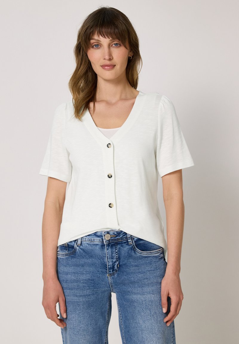 Femme aux cheveux bruns mi-longs portant un haut blanc à manches courtes boutonné et un jean bleu sur fond uni.