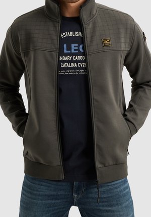 Homme portant une veste matelassée olive foncée ouverte sur un t-shirt graphique noir et un jean bleu, mains dans les poches de la veste.