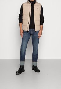 Beige puffer vest med hög krage och logotyp, kombinerad med en svart tröja, mörkblå jeans och svarta ankelboots med tjocka sulor.