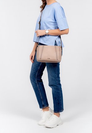 Hnědá crossbody taška s hřebíkovými akcenty, obdélníkový tvar, kožený materiál, nastavitelný popruh, nošena s lehce modrým topem a modrými džíny.