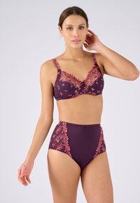 Mujer vestida con un sujetador morado a juego y braguitas de cintura alta con bordados florales en rosa y naranja, posando con la mano cerca de la oreja.