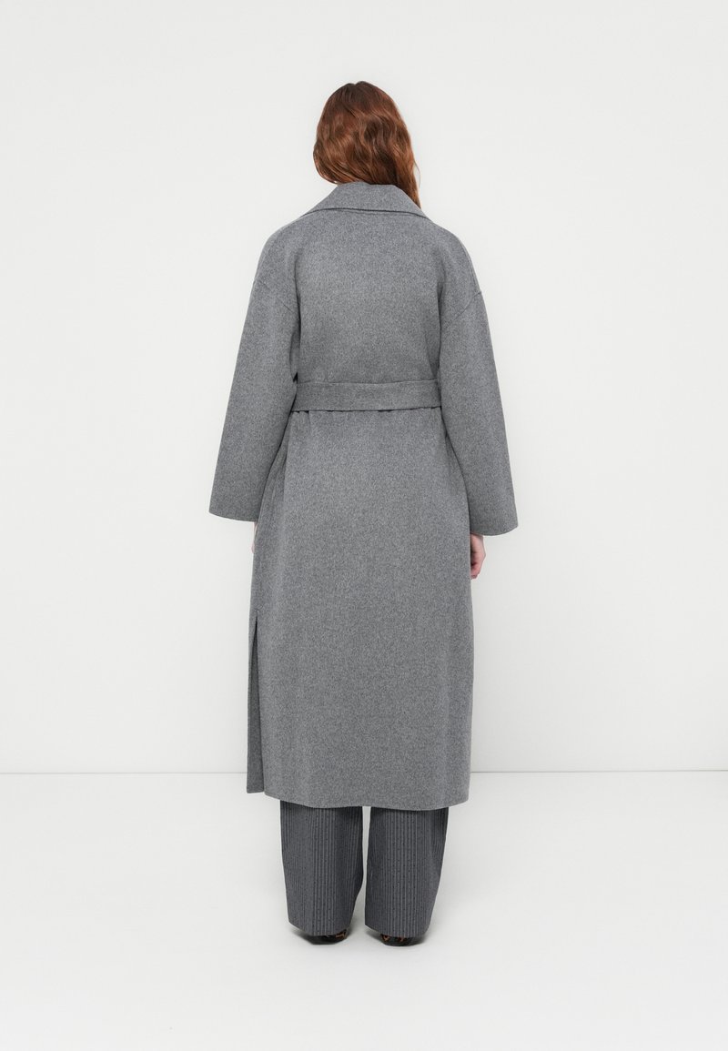 Donna con lunghi capelli che indossa un cappotto grigio con cintura e pantaloni grigi a righe larghe, di fronte a un muro bianco.