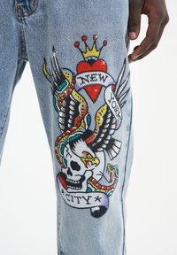 Jeans in denim con un vivace design ricamato che presenta un teschio, ali e un cuore, abbellito con perline colorate e un banner con la scritta "NEW YORK CITY."