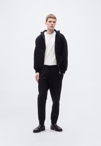 Μαύρο φούτερ με φερμουάρ και μπροστινές τσέπες πάνω από λευκό μπλουζάκι· μαύρο παντελόνι φόρμας και μαύρα μοκασίνια ολοκληρώνουν την εμφάνιση. Απαλή, καθημερινή στιλιστική επιλογή.