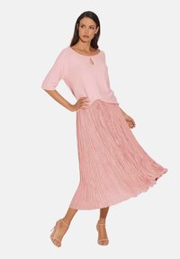 Helles rosa Strickoberteil mit einem Schlüsselaugen-Detail, kombiniert mit einem plissierten Midi-Rock in einem passenden Farbton. Das Ensemble zeichnet sich durch einen lockeren Schnitt und einen fließenden Stoff aus.