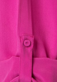 Le tissu de la robe fuchsia présente une texture lisse avec un détail de bouton sur une bretelle. Les coutures sont visibles, renforçant la structure du design.