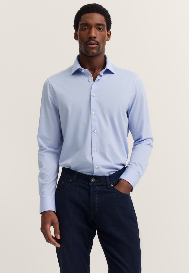 Hellblaues Hemd mit langen Ärmeln und Button-down-Kragen, kombiniert mit dunklen Denim-Jeans. Das Hemd hat eine glatte Textur und einen subtilen Glanz.