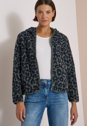 Donna che indossa una giacca pelosa con stampa leopardata blu e verde con zip sopra una camicia bianca e jeans blu, in piedi davanti a uno sfondo chiaro.