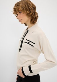Maglione a maglia crema con accenti neri, caratterizzato da un colletto con bottoni e due tasche sul petto bordate di nero. Maniche lunghe con polsini neri.