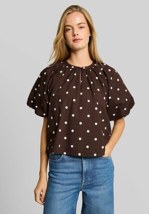 Donna che indossa una camicetta marrone a maniche corte con pois bianchi e jeans blu, in piedi con una mano in tasca su uno sfondo semplice.