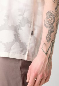 Cămașă ușoară și transparentă, de culoare albă murdară, cu un model floral, având un etichetă laterală. Pantaloni bej cu o textură netedă și croială ajustată.