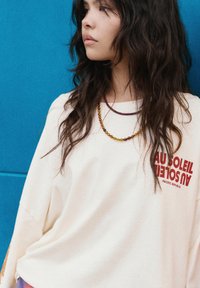 Cremfarbener Oversized-Pullover mit rotem Grafikkreistext, kombiniert mit mehrfarbigen Perlenhalsschmuck. Der Hintergrund ist in einfarbigem Blau.