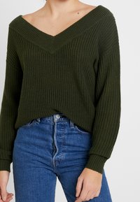 Mörkgrön stickad tröja med djup V-ringning och långa ärmar, med ribbad struktur. Kombinerad med högmidjade blå jeans.