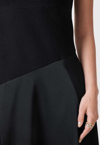 Abito nero con un corpino a coste testurizzate e una gonna liscia, con un design a strati. È anche visibile una mano adornata con anelli.