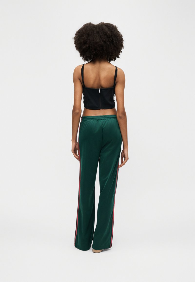 Femme aux cheveux bouclés portant un crop top noir avec une fermeture éclair au dos et un pantalon large vert avec des bandes rouges sur les côtés, debout de dos.
