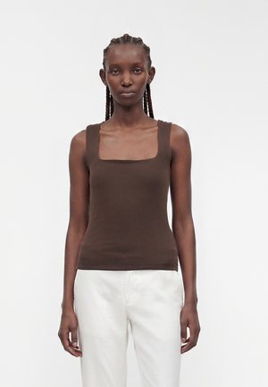 Donna con i capelli intrecciati che indossa un top marrone senza maniche e pantaloni bianchi, in piedi davanti a uno sfondo chiaro e uniforme.
