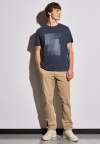 T-shirt bleu marine à manches courtes avec un graphique géométrique dans des teintes plus claires, associé à un pantalon khaki et des baskets de couleur claire.