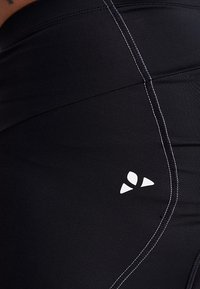 Leggings de sport noirs avec une texture lisse, dotés de coutures blanches contrastantes et d'un petit logo sur le côté. Vue en gros plan.