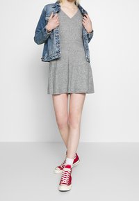 Robe en tricot grise avec un décolleté en V, jupe évasée, associée à une veste en jean. Le modèle porte des baskets montantes rouges et des chaussettes blanches.