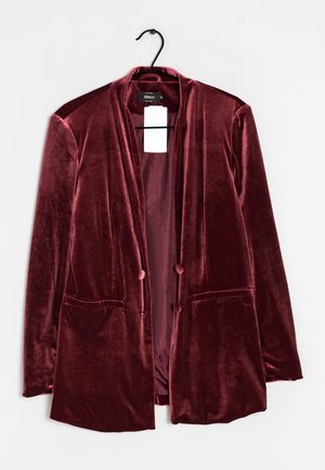 Fluwelen bordeauxblazer met twee voorzakken en met stof overtrokken knopen, hangend aan een zwarte hanger met een wit prijskaartje binnenin de kraag.