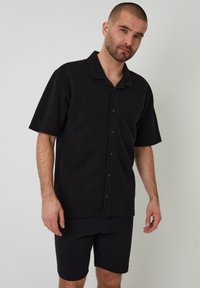 Schwarzes Kurzarm-Hemd mit Knöpfen und Kragen, aus strukturiertem Stoff, kombiniert mit passenden schwarzen knielangen Shorts. Einfaches, zeitgemäßes Design.