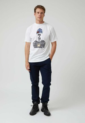 Camiseta blanca de manga corta con un gráfico de una figura que lleva un gorro azul y una camiseta a rayas, combinada con pantalones cargo oscuros y botas negras.